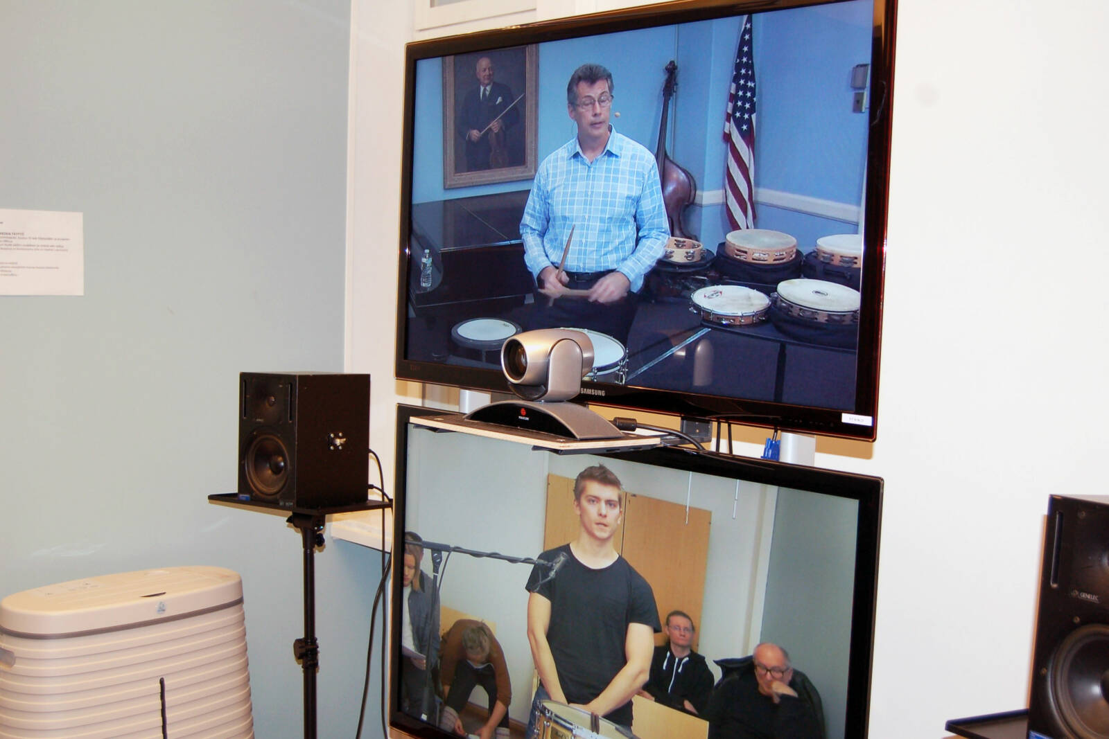 Kaksi näyttöä, joissa toisessa näkyy opiskelija ja toisessa opettaja lyömäsoitinten äärellä.