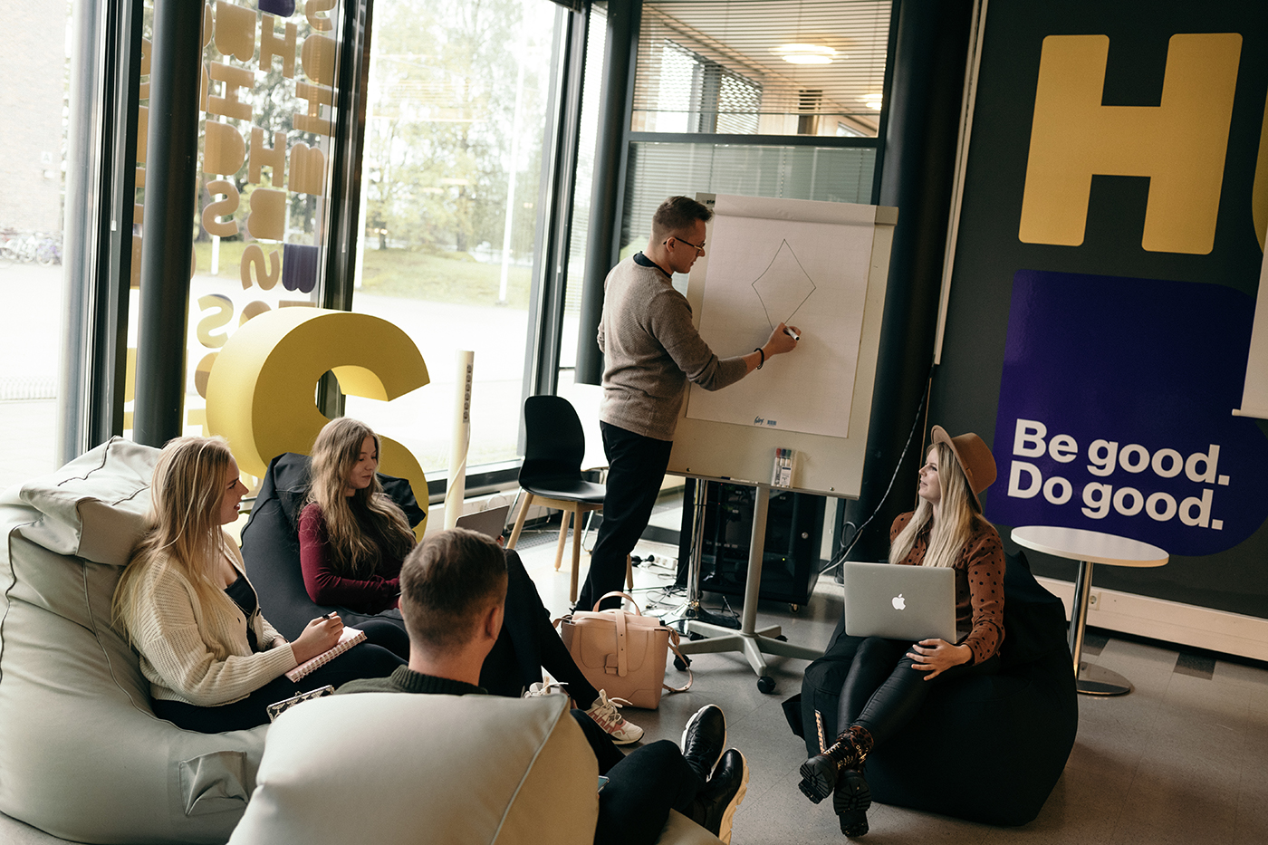 Rento opiskelutilanne, iloiset opiskelijat istuvat säkkituoleilla. Taustalla seinässä lukee Be good - do good.