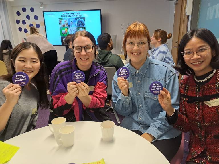 Neljä naista istuu pöydän ympärillä ja pitelevät käsissään rintamerkkiä, jossa lukee "Puhu minulle suomea".