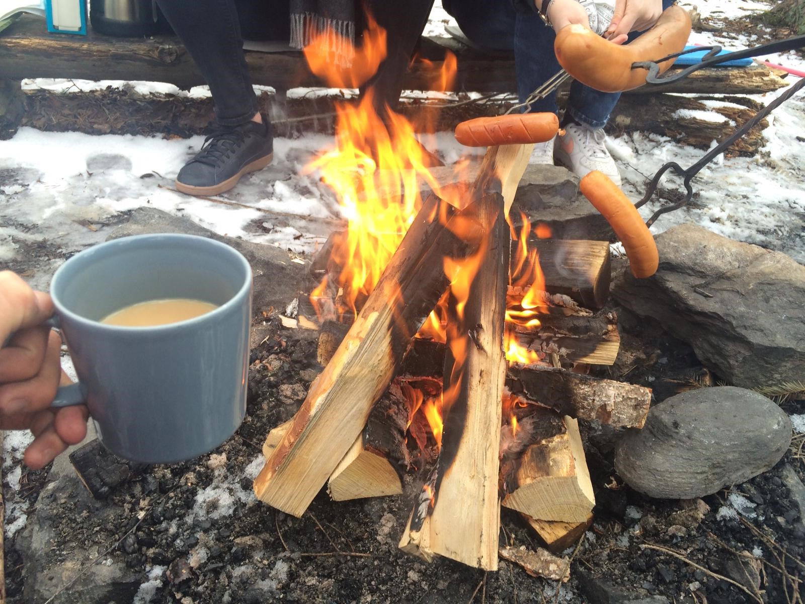 Nuotio, jonka yllä grillataan makkaraa. Etualalla kahvikuppi