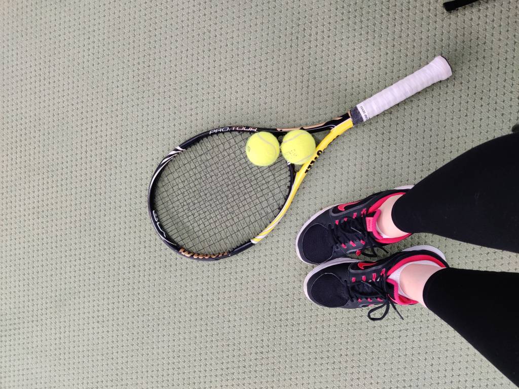 Ylhäältä otettu kuva, jossa näkyy kuvaajan jalat, tennismaila ja kaksi tennispalloa.