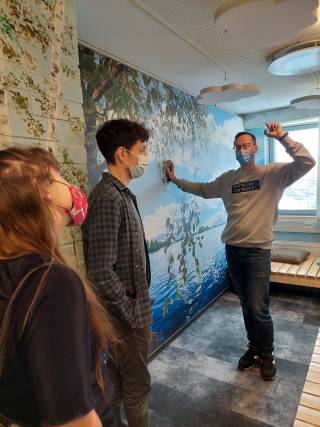 Kuvassa Tulliportin normaalikoulun tilassa, jossa seinämaalaus ja laituri sekä toimijoita kuuntelemassa esittelyä