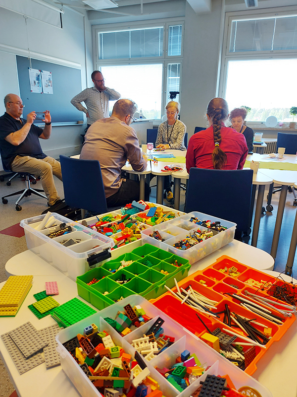 Etualalla erilaisia legoja laatikoissa, taustalla rakenteleva tiimi.