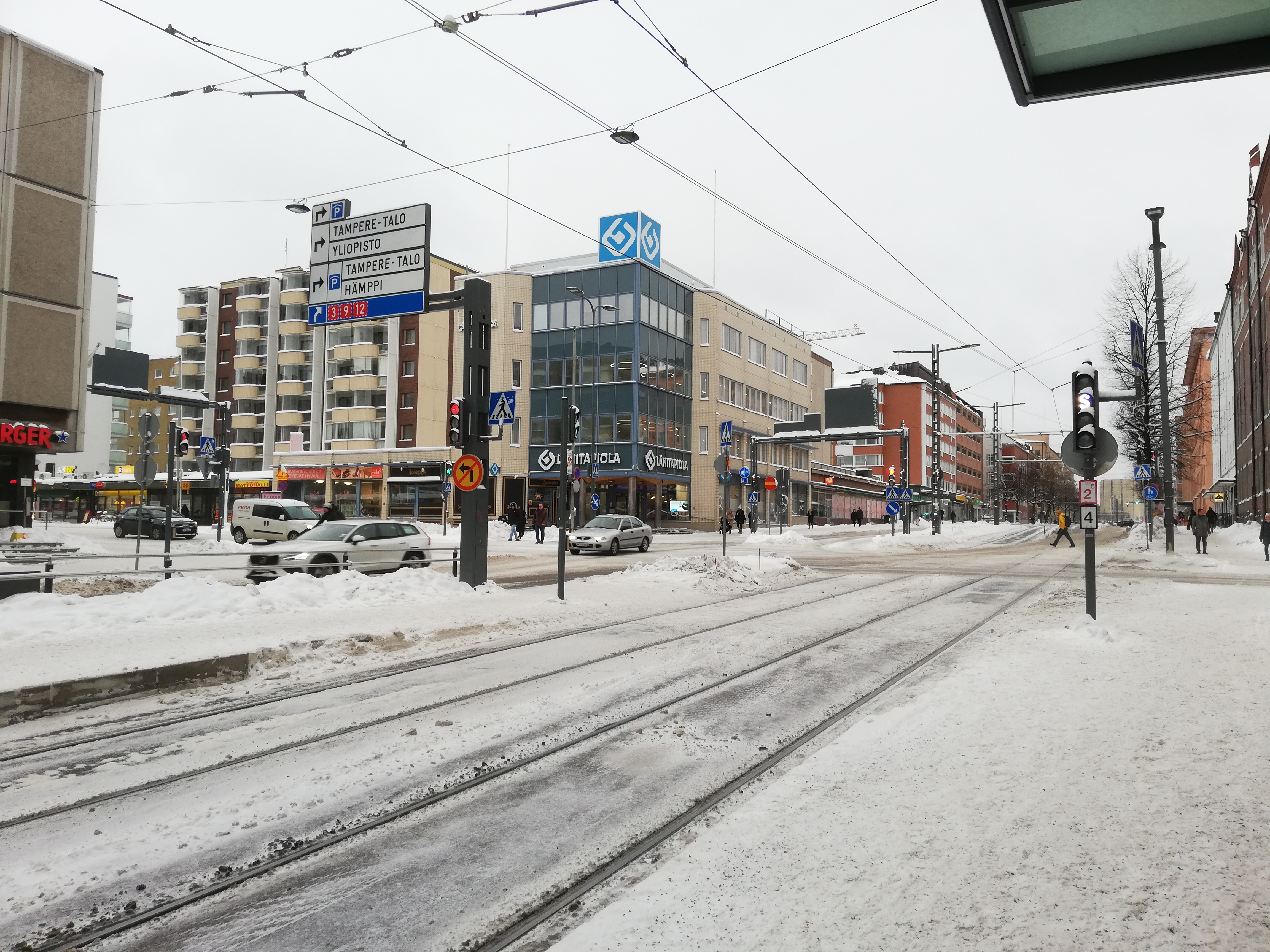 Näkymä Tullin ratikkapysäkiltä. Kuvassa näkyvät raiteet, johdinlankoja, liikennevalot sekä rakennuksia, mm. Lähi-Tapiola Pirkanmaan konttori.