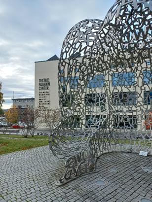 A statue made of metal in front of the Textile Fashion Center building.
