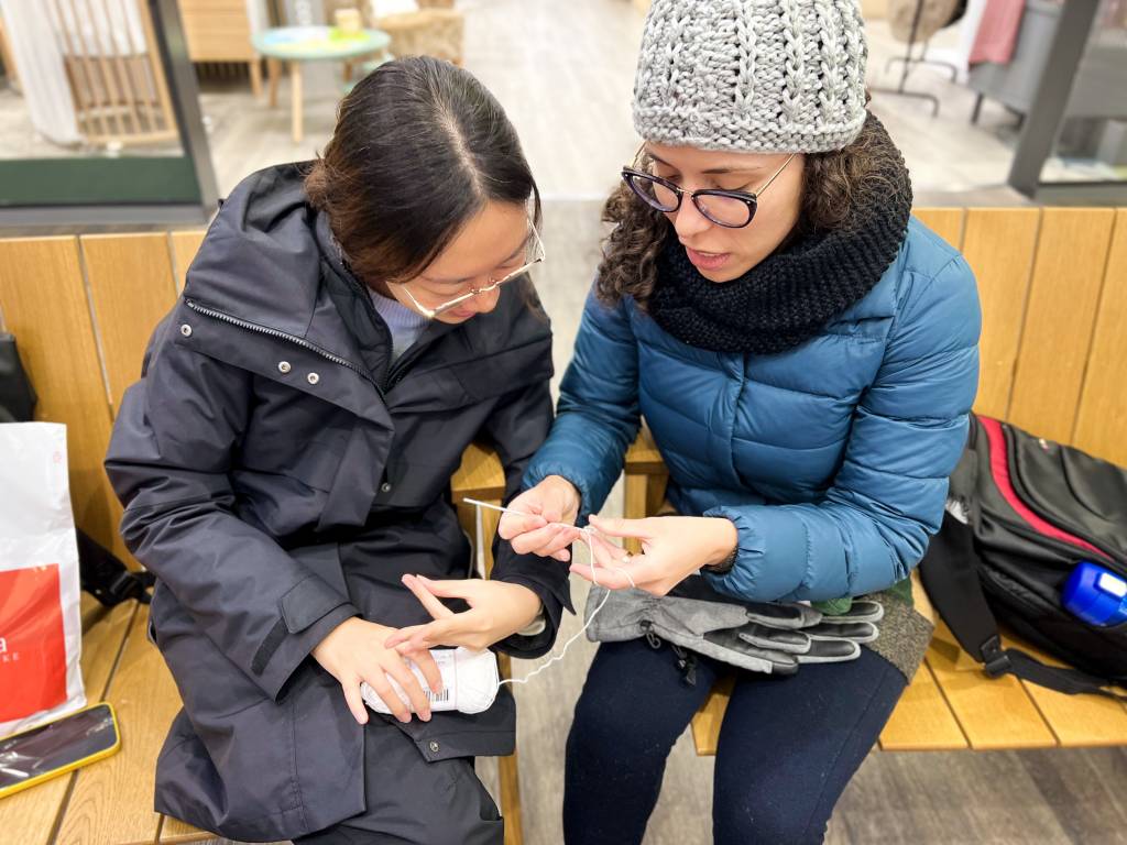 Two people sitting on benches with winter jackets on. The other one is teaching to crochet for the other.