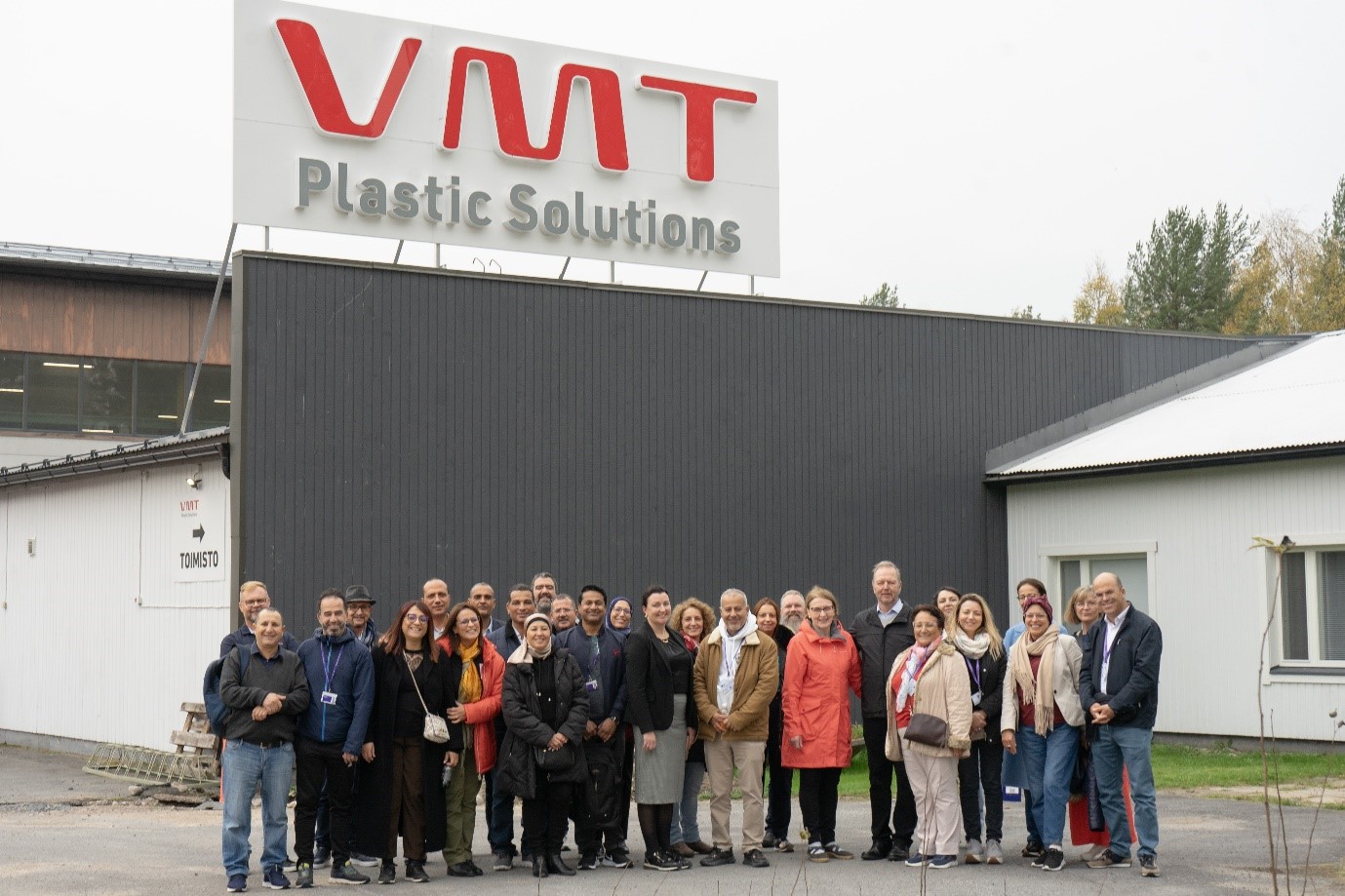 Ihmisiä seisomassa rakennuksen edessä jonka yläpuolella on kyltti, jossa lukee "VMT Plastic Solutions".
