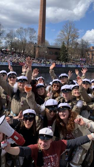 Iloisia ja innostuneita opiskelijoita samppanjankultaisissa haalareissa ja teekkarilakit päässä kosken rannalla