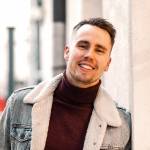 Hymyilevä nuori mies nojaa tiiliseinään farkkutakissa.