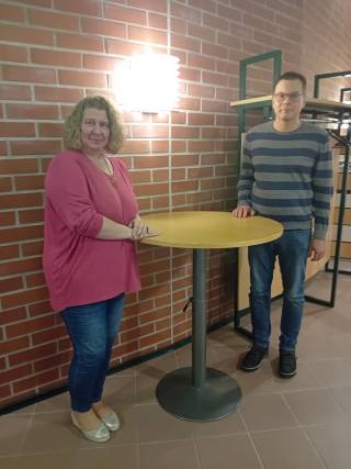 Hankkeen projektipäällikkö ja asiantuntija seivovat hymyillen pyöreän pystypöydän takana tiiliseinän edessä.