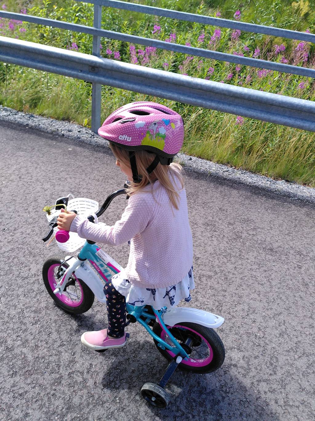 Pieni lapsi pyöräilemässä kesällä apupyörillä varustetulla polkupyörällä. Lapsella vaaleanpunainen villatakki ja kypärä, sekä kädessään voikukka.