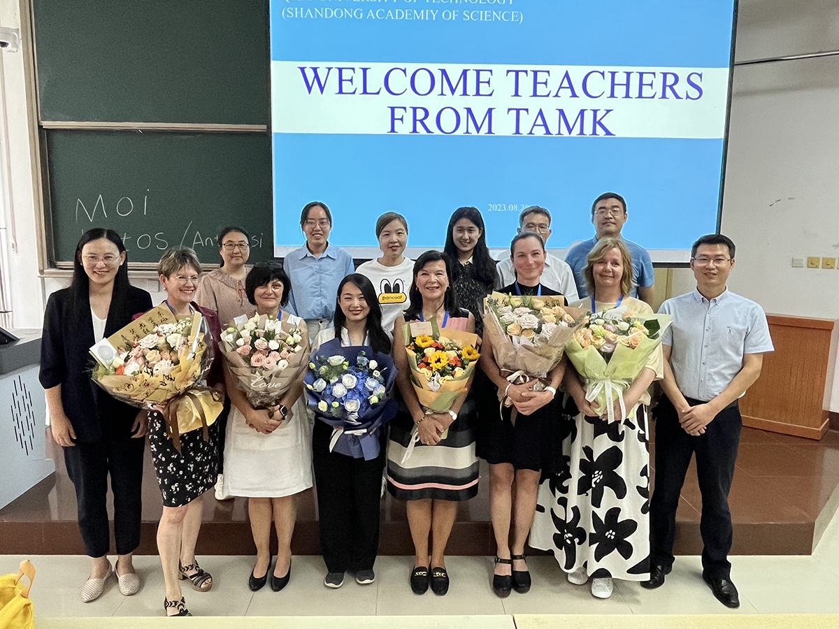 Fourteen people standing in a group with "Welcome teachers from TAMK" text on a screen behind them.