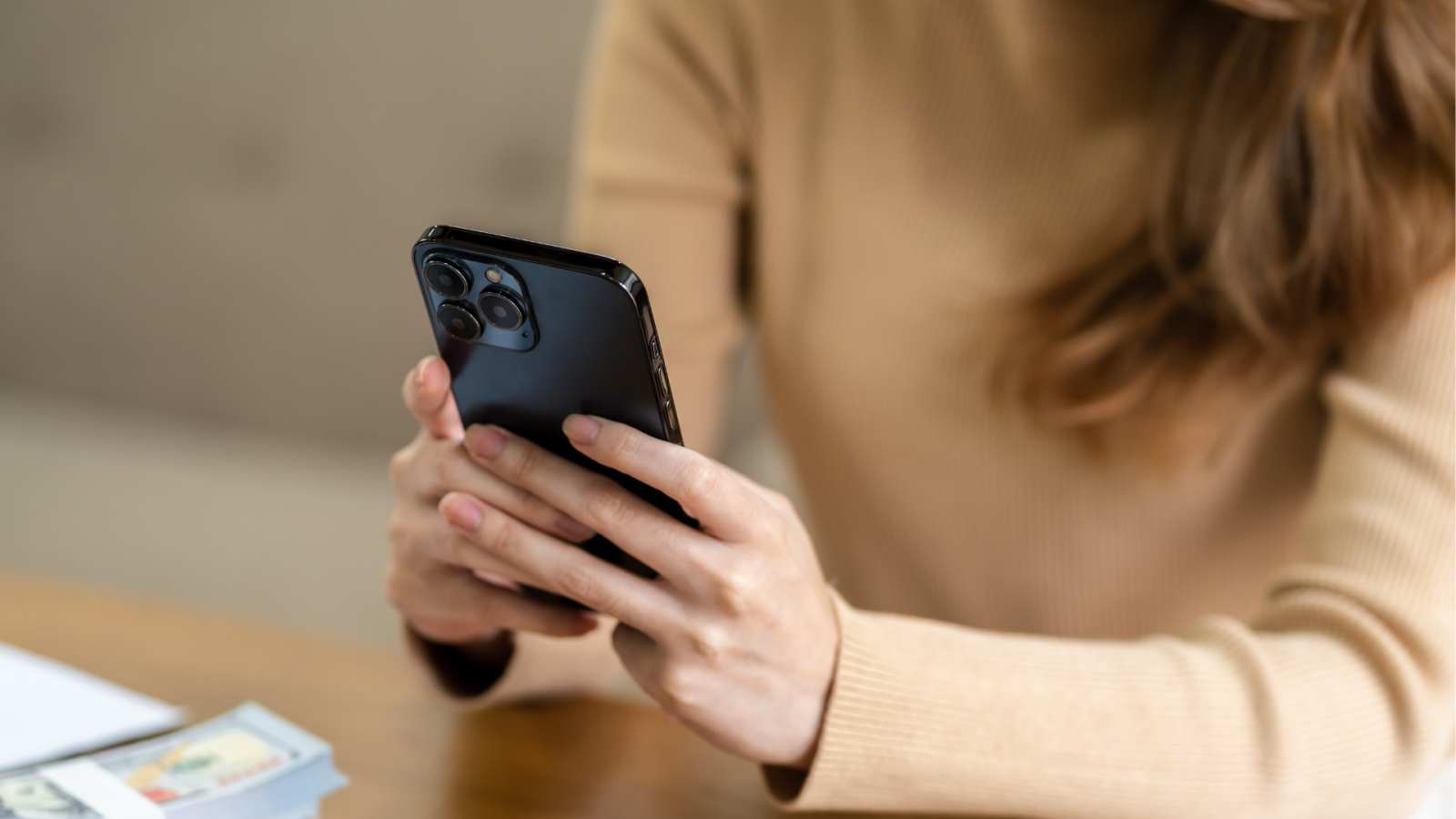 Ihminen pitelee puhelinta istuessaan pöydän ääressä. Henkilön kasvot eivät ole näkyvissä.