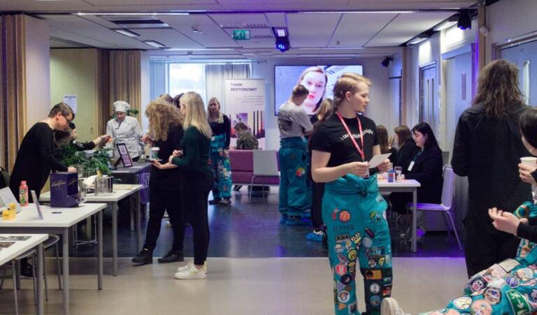 Opiskelija kävelemässä aulassa opiskelijan haalari yllään ja hänen takanaan on ihmisiä, jotka kiertelevät ja katselevat esittelypöytiä.