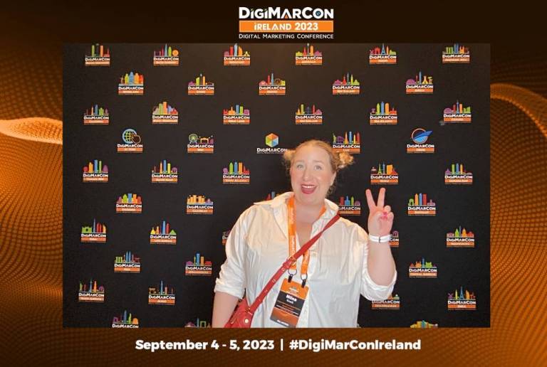 Henkilö näyttää voitonmerkkiä konferenssin kuvausseinän edessä.