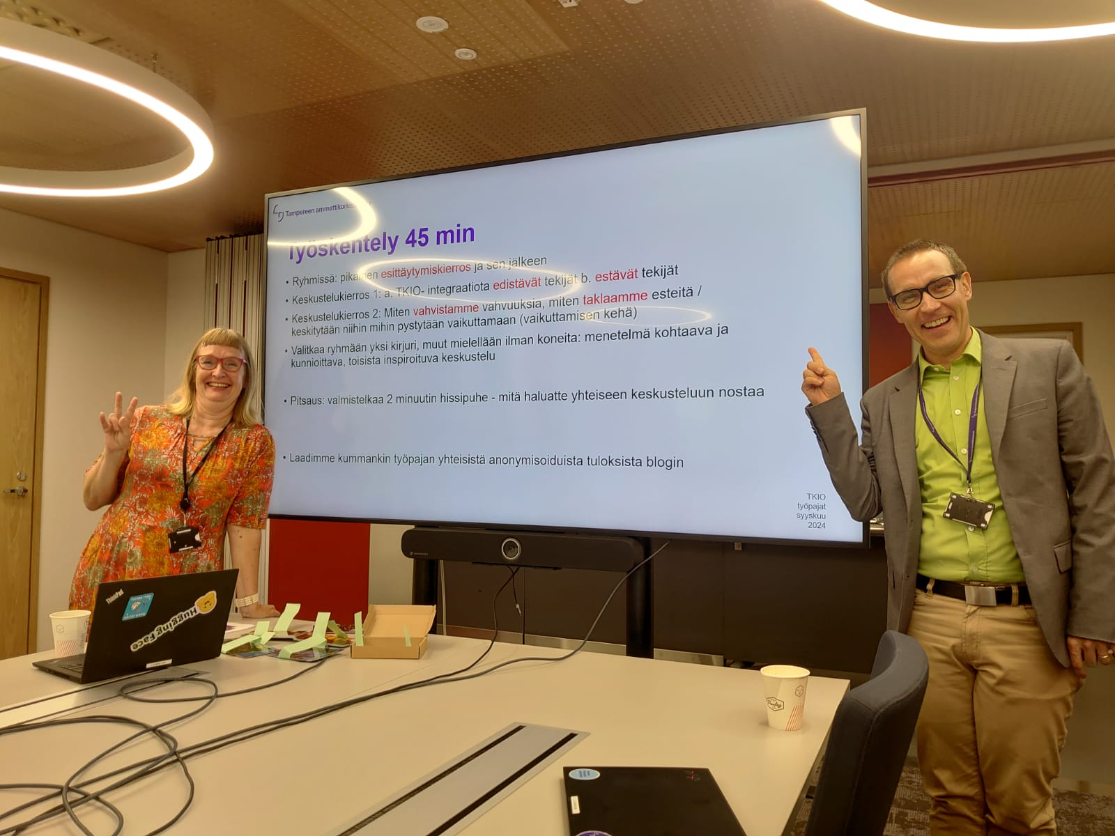 kaksi henkilöä hymyilee ison screenin edessä