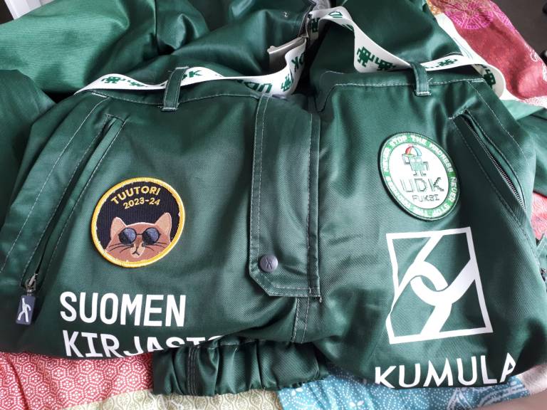 A green student overall with two patches, one is saying tuutori 2023-24
