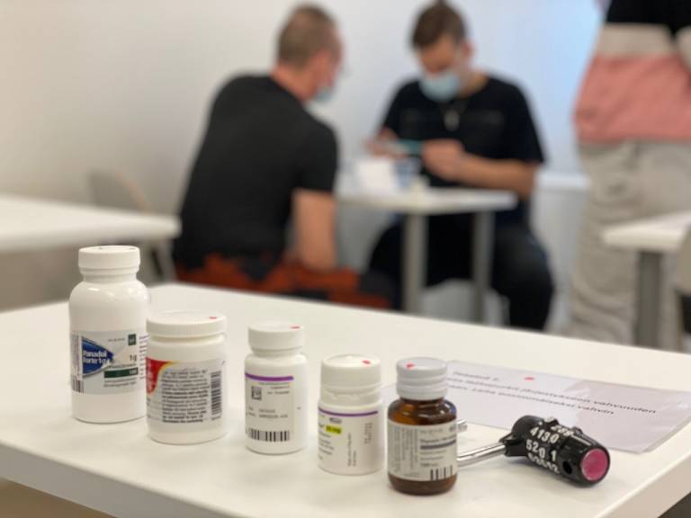 Etualalla erilaisia lääkepurkkejä ja lukko, taustalla pari opiskelijaa laskemassa tehtäviä.