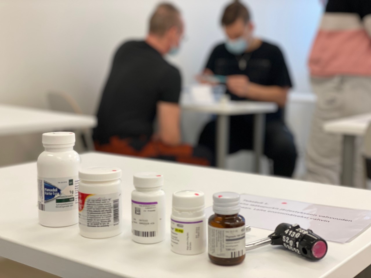 Etualalla erilaisia lääkepurkkejä ja lukko, taustalla pari opiskelijaa laskemassa tehtäviä.