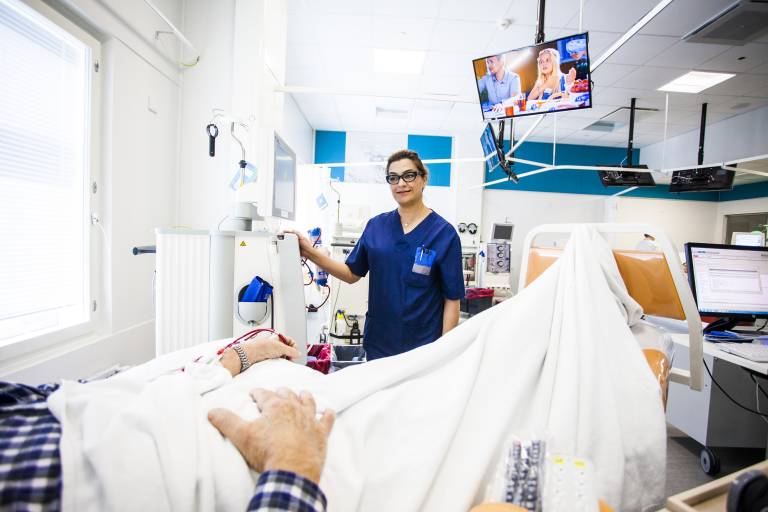 Hoitaja seisoo potilaan sängyn vierellä sinisessä hoitoasussa.