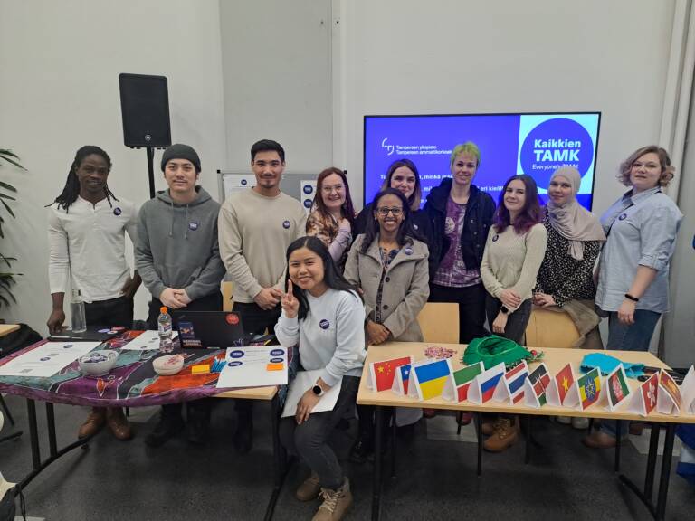 Ihmisiä seisomassa ja istumassa kahden pöydän takana ryhmämuodostelmassa.