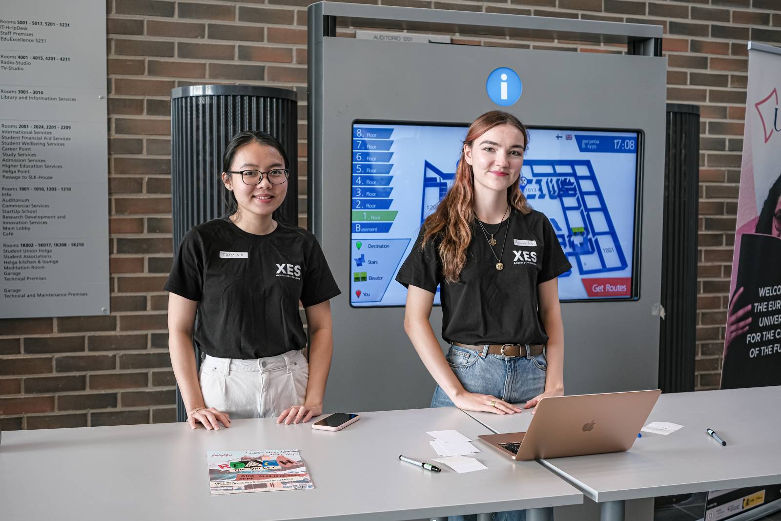 Two students are standing behind the desk. They are wearing associations T-shirts.