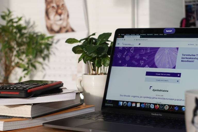 Kuva, jossa tietokoneella auki Tampereen yliopiston www-sivu, kirjapino, viherkasveja ja seinällä kalenteri, jossa eläin