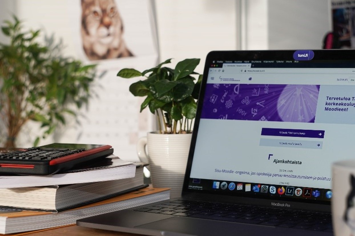 Kuva, jossa tietokoneella auki Tampereen yliopiston www-sivu, kirjapino, viherkasveja ja seinällä kalenteri, jossa eläin