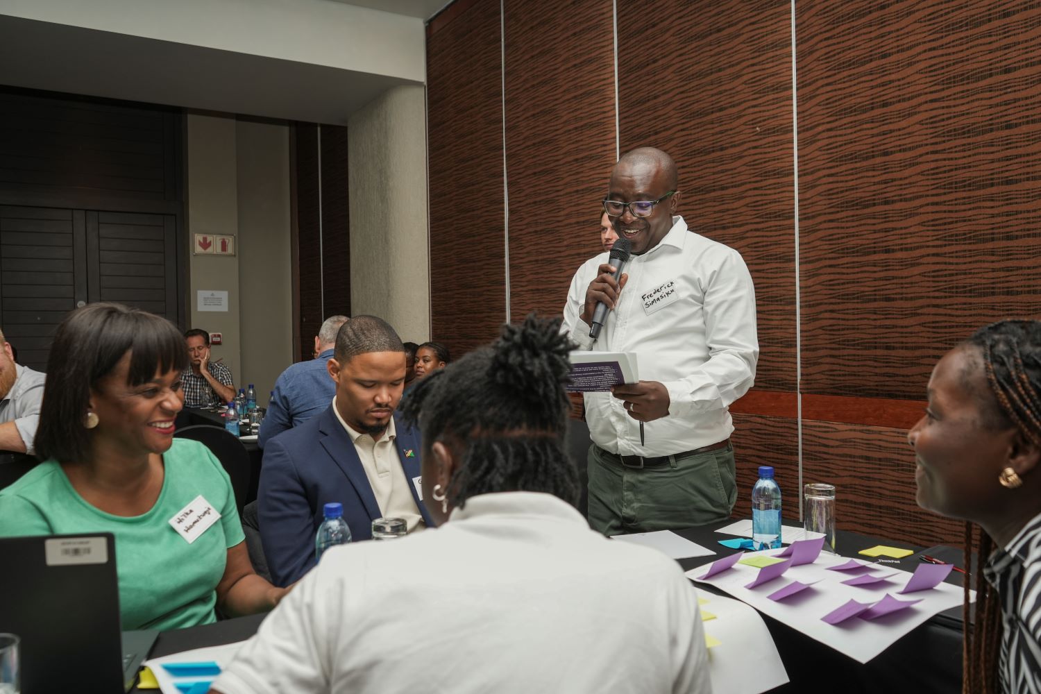 Ihmisiä istumassa pöydän ympärillä ja kuuntelemassa miestä, joka seisoo ja puhuu.