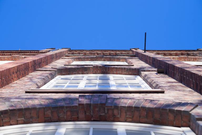 Katsot kuvassa ylöspäin kohti sinistä taivasta punaisen tiiliseinän edessä. Tiiliseinässä on valkoiset ruutuikkunat.