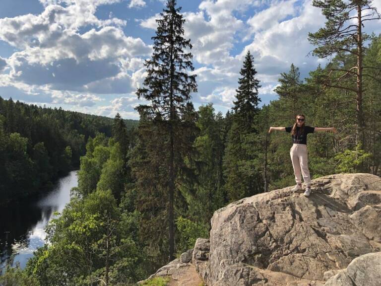 A picture with the nature landscape and the young woman standing on the boulder. She is looking at the camera and smiling.