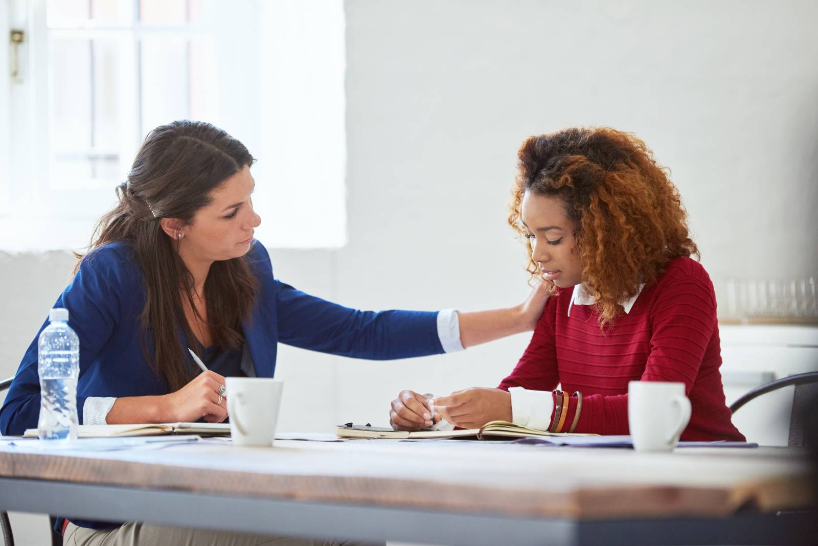 Two women sitting around the table and the other woman has put her hand on other ones shoulder.