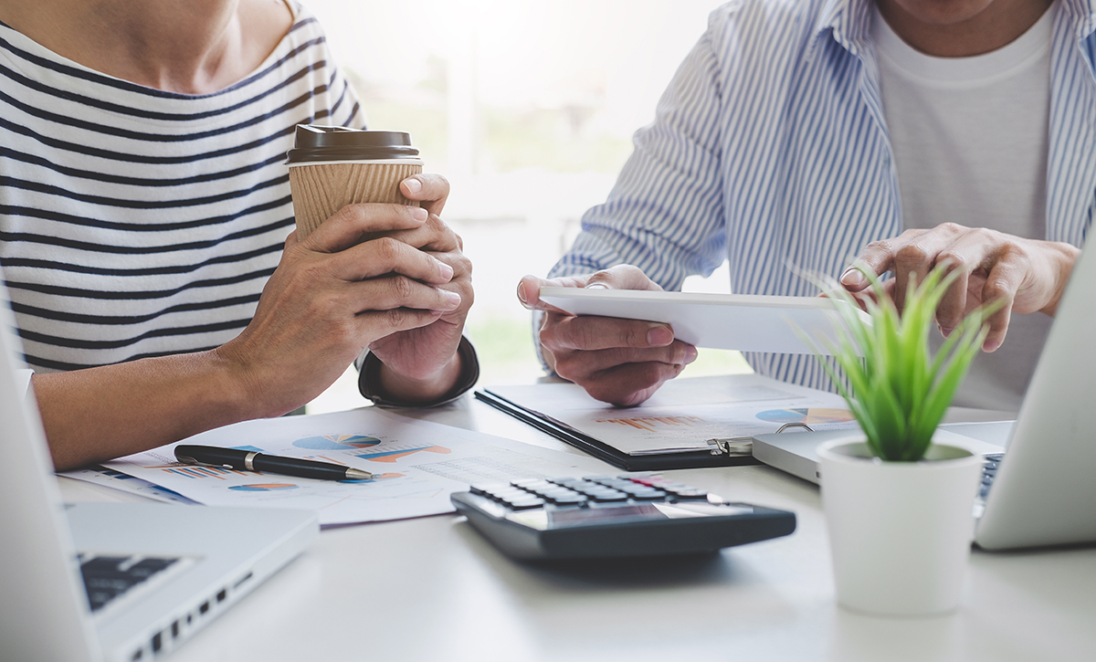Kahden henkilön liikeneuvottelu pöydän ääressä, pöydällä tietokone, laskin ja papereita.