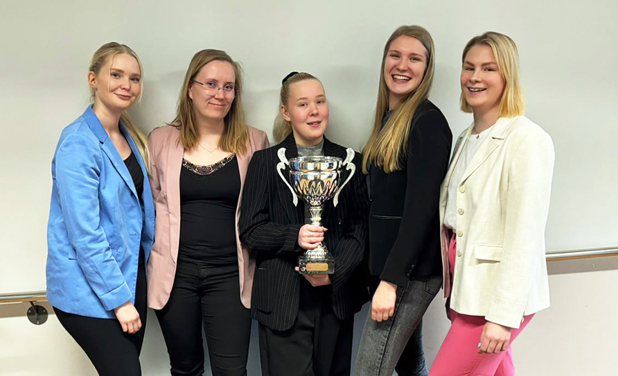 Viisi nuorta naista käytävällä pokaali kädessä.