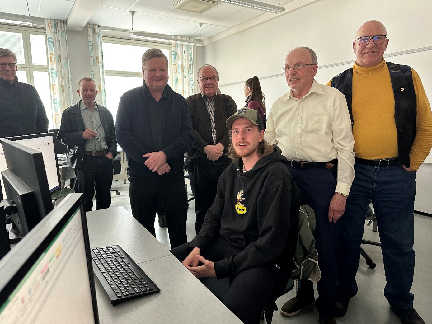 Mies istumassa tietokoneen äärellä ja häntä ympäröiden seisomassa seitsemän aikuista.