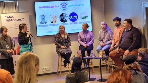 Ihmisiä istumassa lavalla tuoleissa ja heidän vieressään seisoo juontaja, joka pitää mikrofonia kädessään.