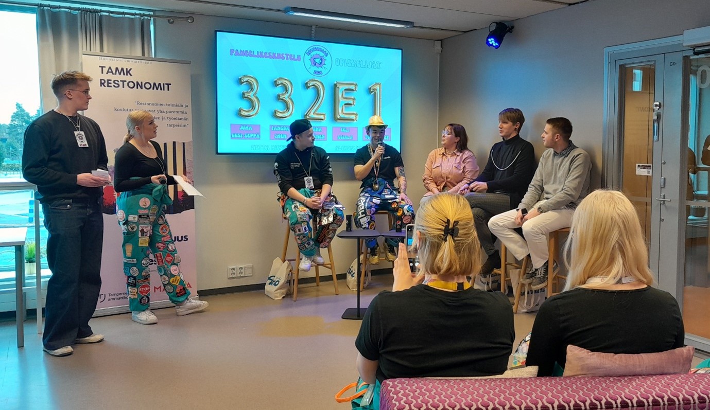 Viisi ihmistä istumassa lavalla, joista yksi puhuu mikrofoniin. yleisön katsellessa.