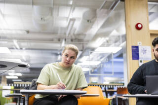 Kaksi opiskelijaa istumassa luokkahuoneessa. Toisen edessä on kone ja toinen tekee muistiinpanoja tabletille.