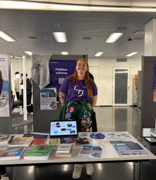 Claudia messupöydän takana, päällä opiskelijalähettiläspaita ja opiskelijahaalarit.