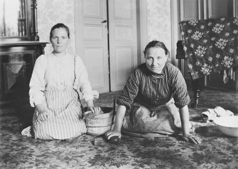 Mustavalkoinen valokuva kahdesta naisesta pesemässä hienon asunnon lattiaa.