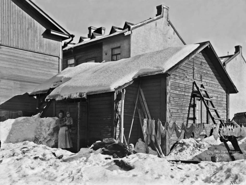 Talvinen valokuva takapihalta 1900-luvun alun Helsingissä, kuvassa nainen, lapsia ja pyykkejä narulla.